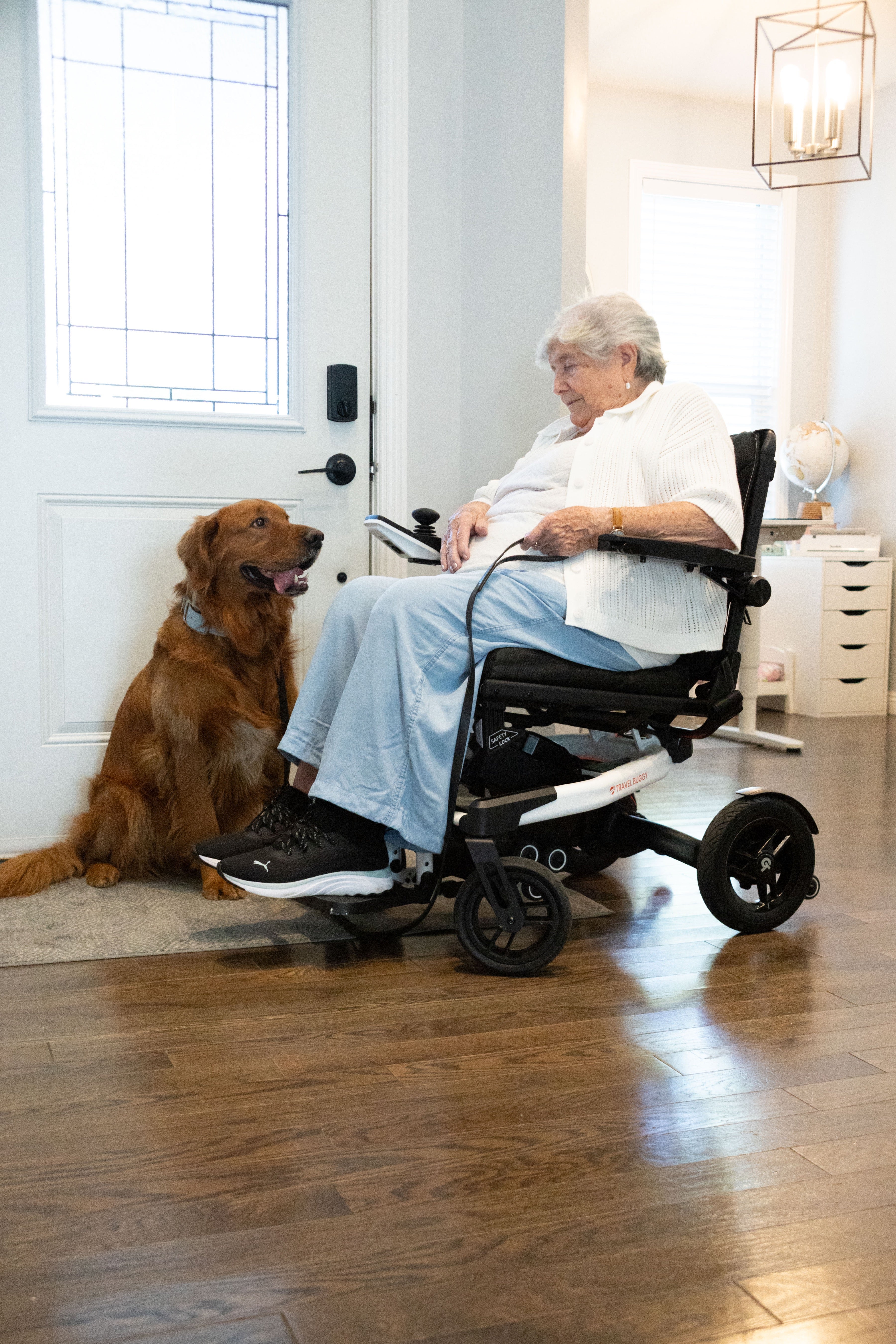 Woman seated in a white Travel Buggy Grand Tourer GT power wheelchair beside a dog at home